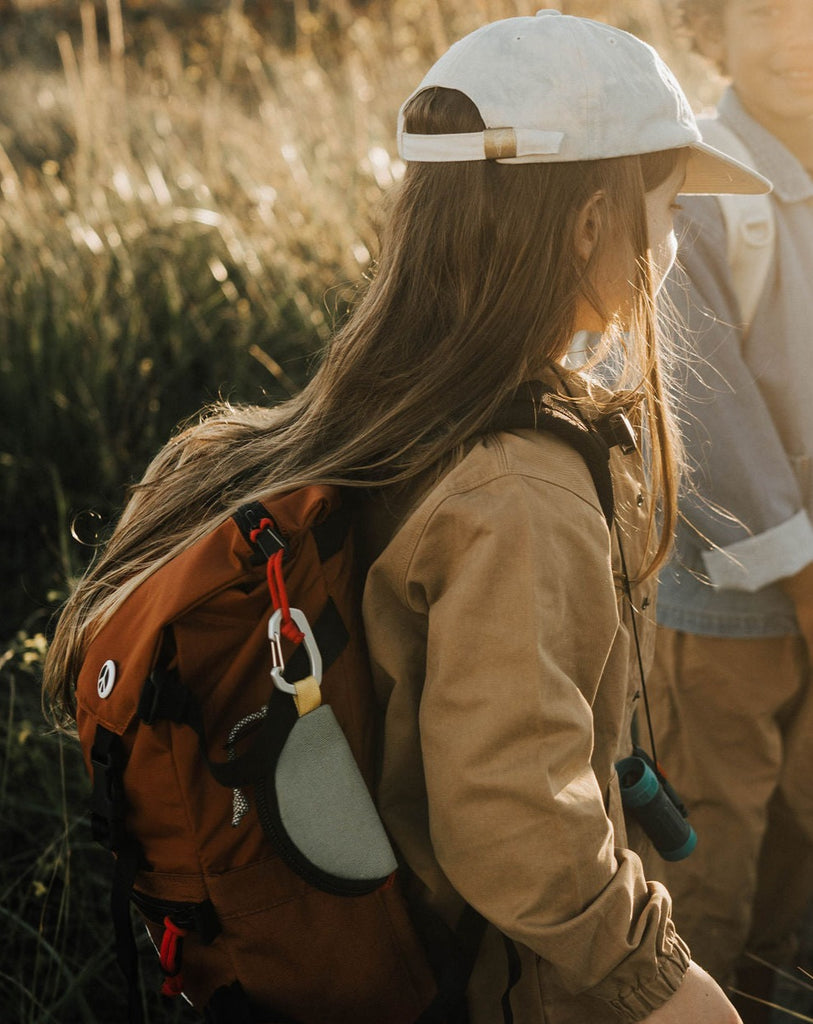 Child with long hair and white baseball cap standing in a field at golden hour wearing the Topo Designs Rover Pack Classic in Clay Nylon with Topo Designs Taco Bag in Gray attached. 