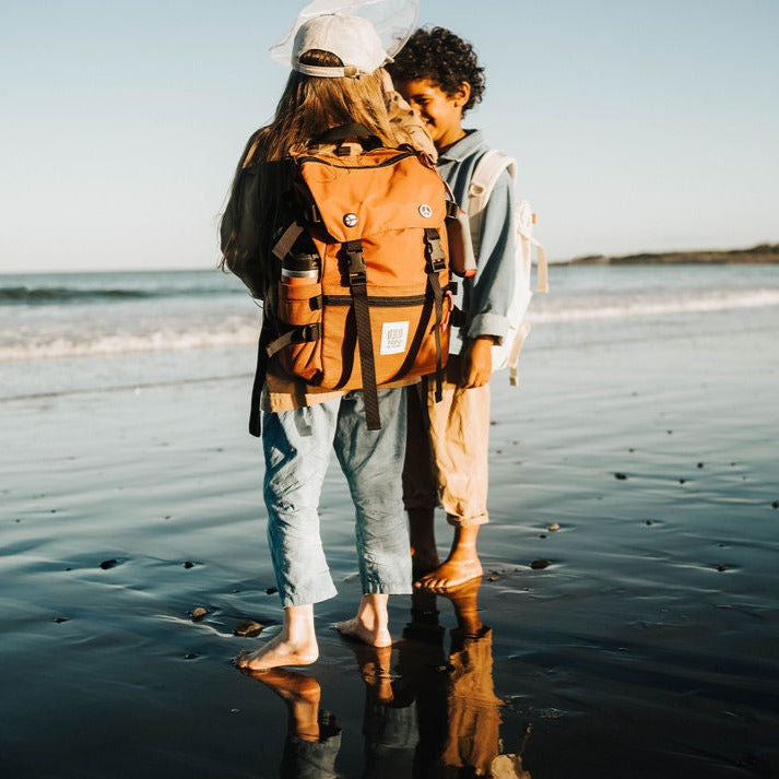 Two children at the beach during golden hour, standing barefoot in the wear and laughing together. One child is wearing the Topo Designs Rover Pack Classic in Clay nylon with water bottle and pins attached. The other child is wearing the Topo Designs Rover Pack Classic in Natural Canvas. 