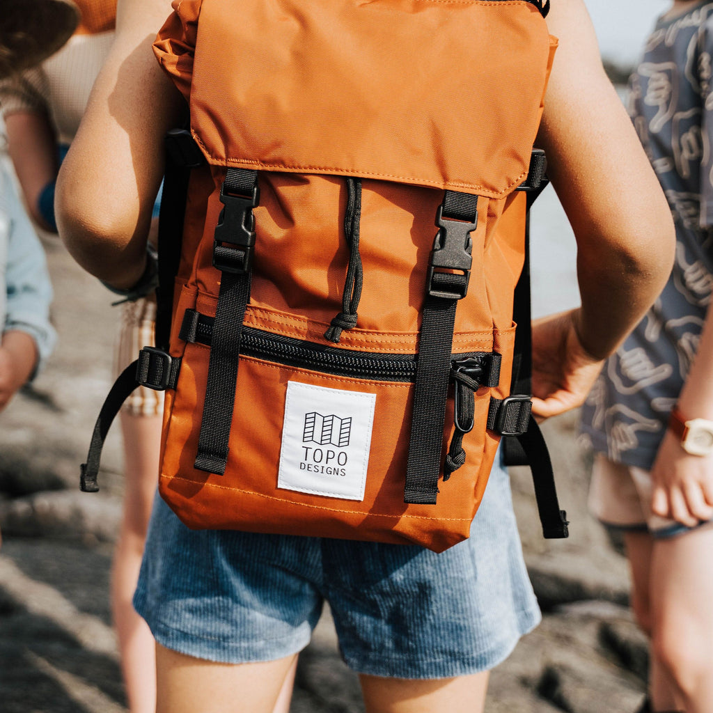 Child wearing a Topo Designs Mini Rover backpack in Clay nylon with black nylon straps, black buckles, black zippers and white and black Topo Designs label on the front pocket. Child has back to camera and is standing with a few other children on the rocks at a beach. 