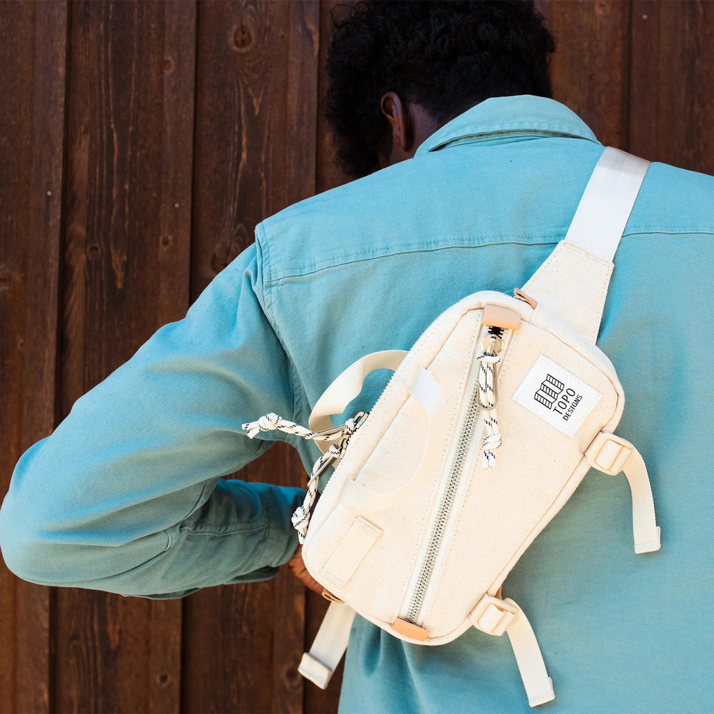Topo Designs Mini Quick Pack in Natural Canvas shown on the back of a man wearing a blue workwear jacket. The man is looking down and facing a distressed wooden wall. 