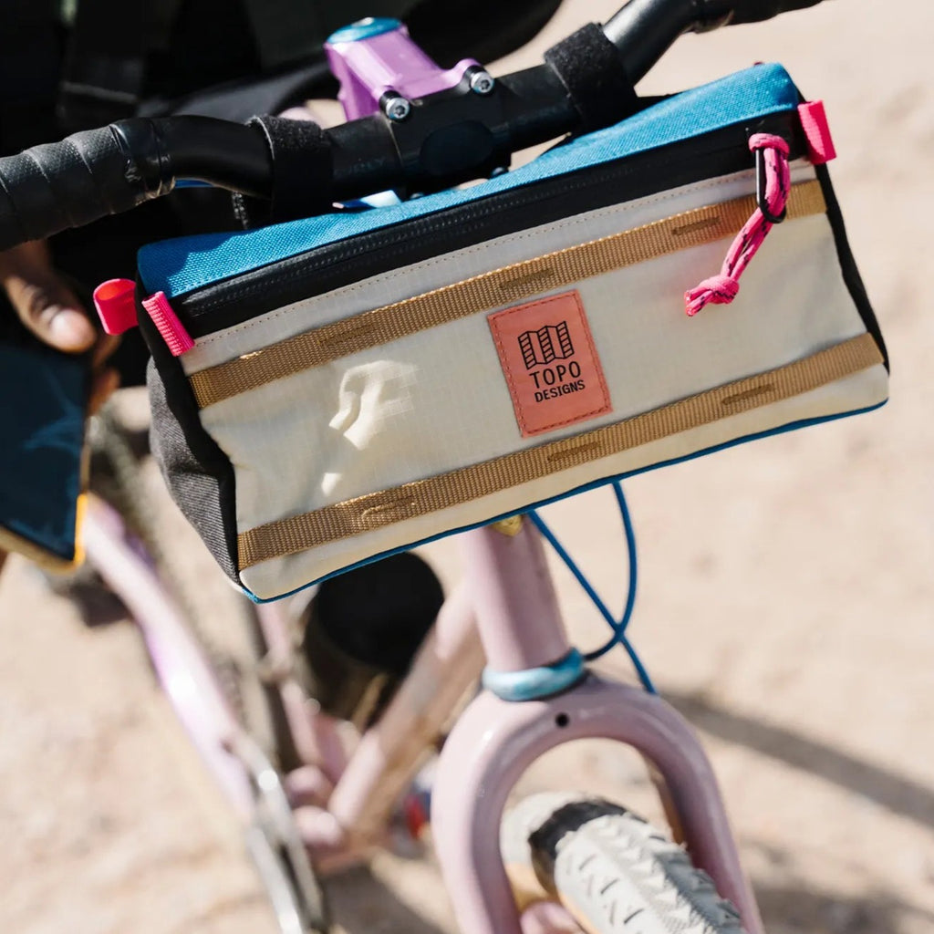 Topo Designs Bike Bag Mountain in Bone White/Blue shown attached to the handlebars of a pink mountain bike. 